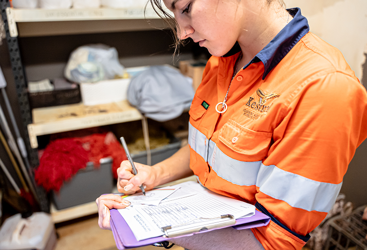 Kestrel Cleaning team member in high-vis shirt filling out safety checklist on clipboard in supply room